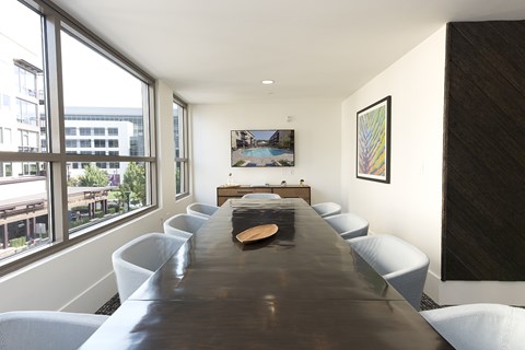 A long brown table is surrounded by white chairs in a room with a large window.