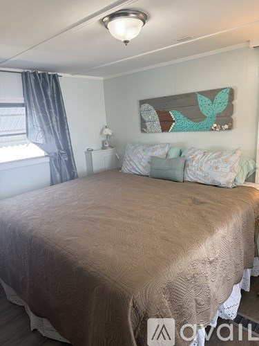 A bedroom with a bed, pillows, a nightstand, a window with curtains, and a wooden piece of artwork on the wall.