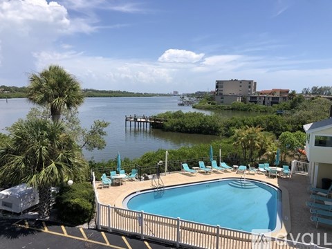 A pool surrounded by chairs and a fence with a view of a body of water and a house in the background.