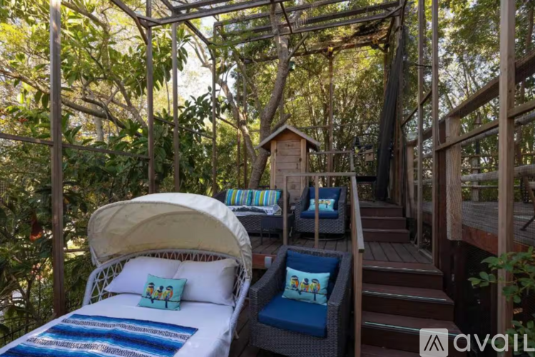 A bed with a white canopy and blue cushions is set up on a wooden deck with a hammock and a small hut in the background.