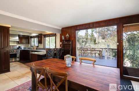 A wooden dining table with chairs is in the middle of a room with a kitchen in the background.