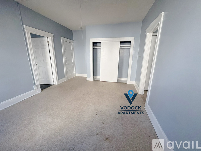 A room with carpeted floor and white walls, with a logo for Vodock Apartments in the bottom right corner.