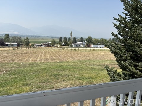 A view from a balcony overlooking a field with a house and trees in the distance.