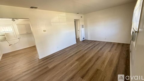 A room with wooden flooring and white walls, with a kitchen area visible in the background.
