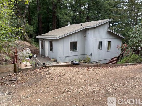 A small white house with a brown roof is surrounded by trees and has a gravel driveway.