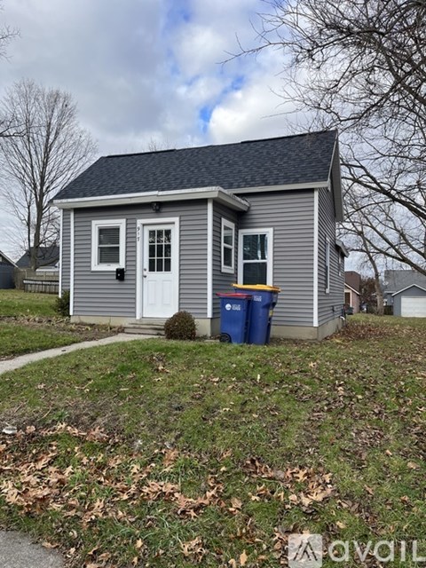 A small grey house with a white door and windows is in front of a blue trash can.