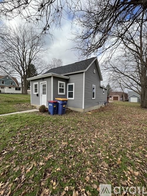 A small house with a grey roof and a blue trash can in front of it.