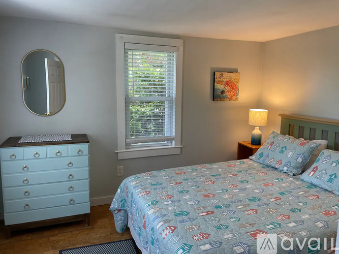 A bedroom with a patterned bedspread, a dresser, and a window with blinds.