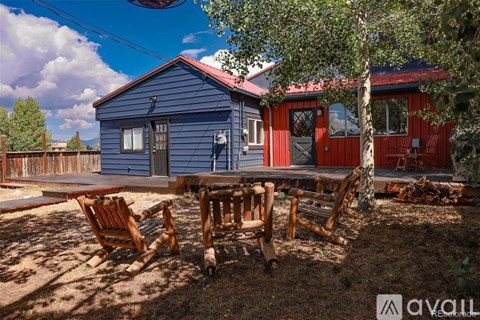 A blue house with a red door and windows is surrounded by trees and has a wooden table and chairs in front.