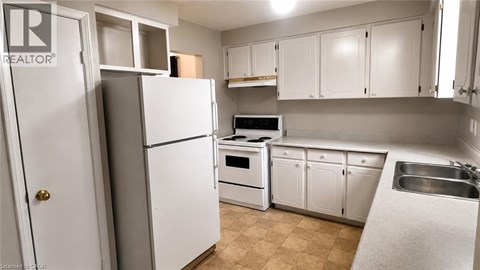 A white refrigerator stands in a kitchen with white cabinets and a stainless steel sink.