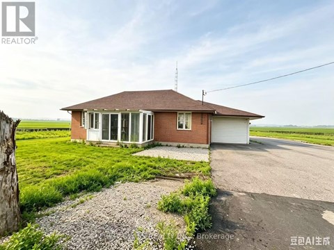 A house with a brown roof and a white garage door is surrounded by a gravel driveway and a grassy field.