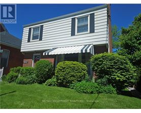 A house with a white siding and a striped awning is surrounded by green bushes.