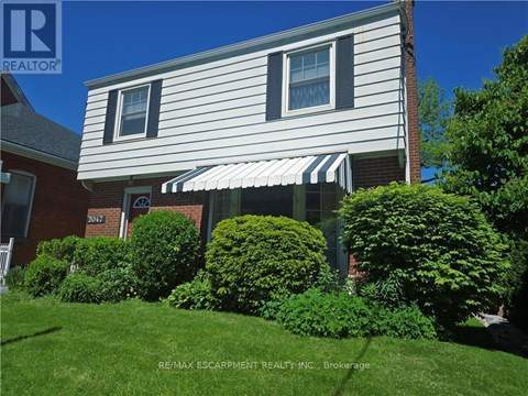 A house with a white siding and a striped awning is surrounded by green bushes.