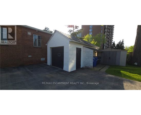 A small white building with a black door is in front of a brick building with a RE/MAX Escarpment Realty Inc. sign.