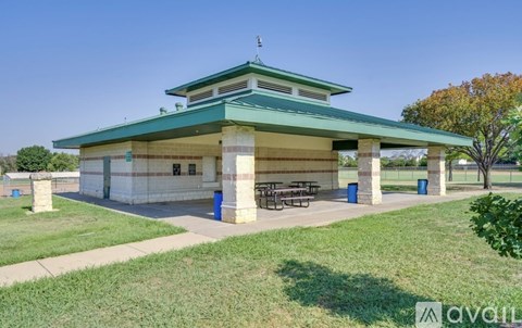 A pavilion with a green roof is surrounded by a grassy area and a clear blue sky.