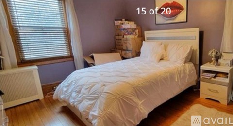A white bed with a white comforter and pillow is in a room with a window and a radiator.