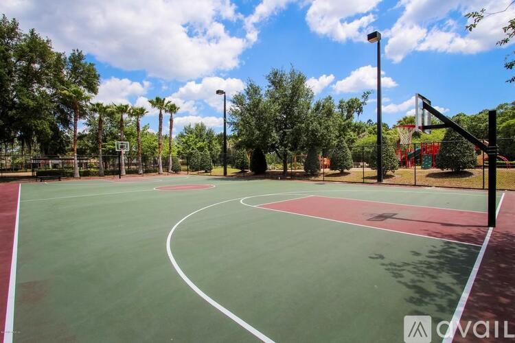 A basketball court with a green surface and white lines, surrounded by trees and a clear blue sky.