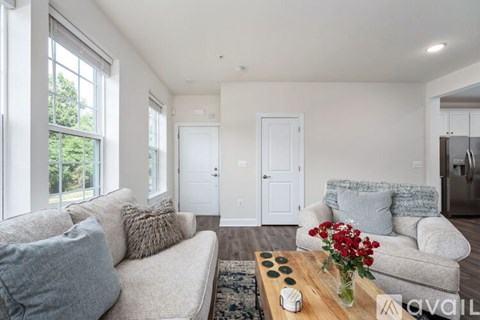 A living room with a grey couch, a coffee table with a vase of red flowers, and a kitchen area in the background.