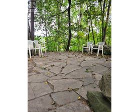 Two white chairs are placed on a stone patio in a wooded area.