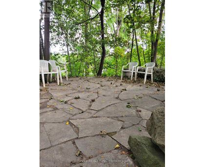 Two white chairs are placed on a stone patio in a wooded area.