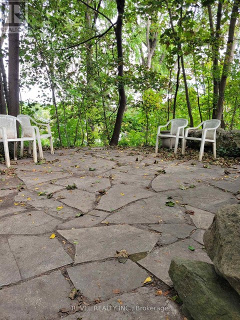 Two white chairs are placed on a stone patio in a wooded area.