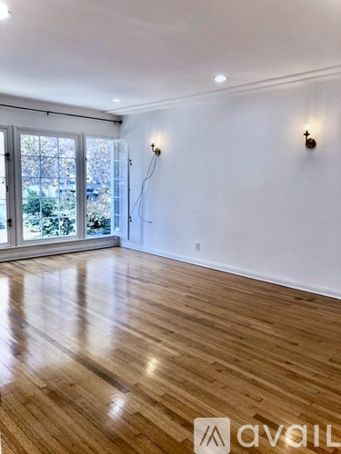 A room with wooden floors and white walls, with two wall-mounted lights and a window with a view of trees and a car.