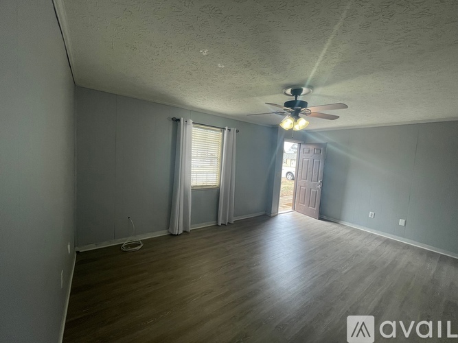 A room with a ceiling fan and light, wooden floors, and a door leading to another room.
