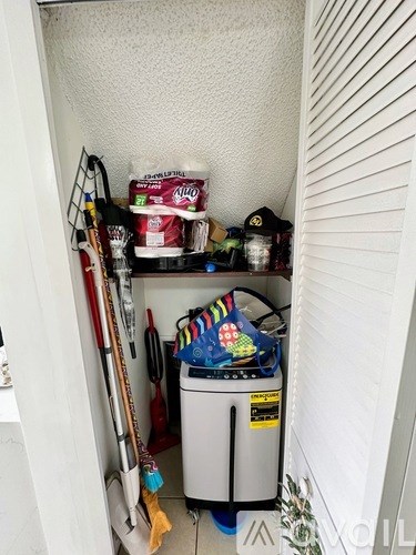 A small storage space with a white wall and contains a variety of items including a red shovel, a white container, and a black bag.