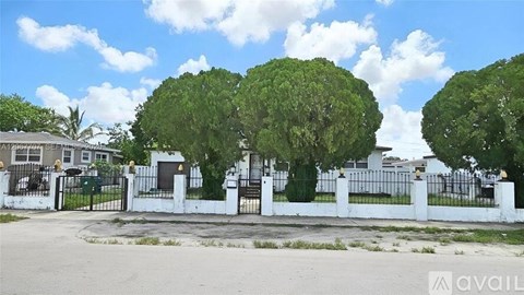 A white fence with a gate stands in front of two large green trees.