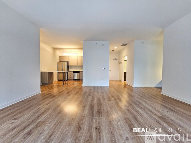 A spacious room with wooden flooring and white walls, with a kitchen area visible in the background.