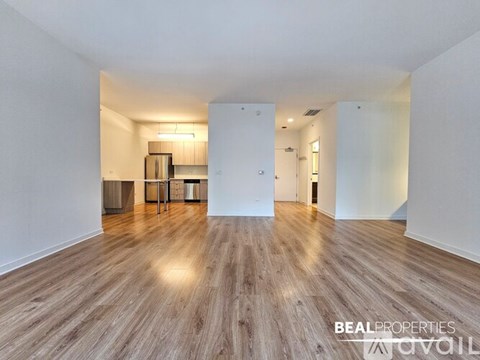 A spacious room with wooden flooring and white walls, with a kitchen area visible in the background.