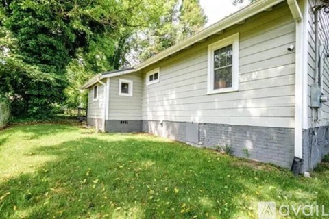 A small house with a grey siding and a white window is surrounded by green grass and trees.
