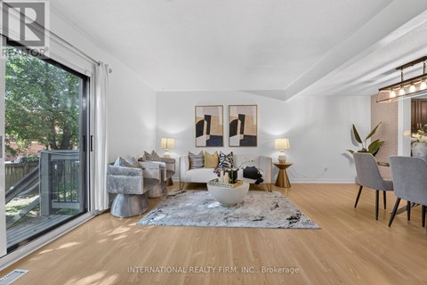 A well-lit, spacious living room with a grey rug, a grey sofa, a white armchair, a grey chair, a wooden floor, a glass door, a table with a vase, a potted plant,