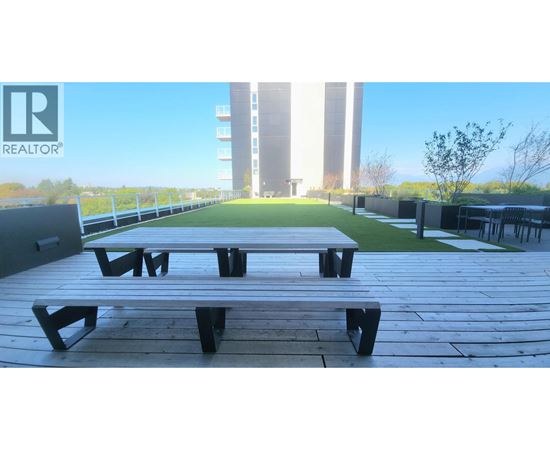 A wooden picnic table is in the foreground of a sunny outdoor area with a building in the background.