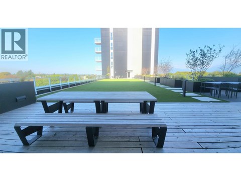 A wooden picnic table is in the foreground of a sunny outdoor area with a building in the background.