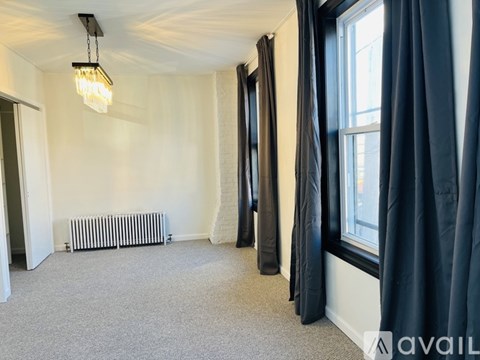 A room with a carpeted floor, a window with black curtains, and a radiator.