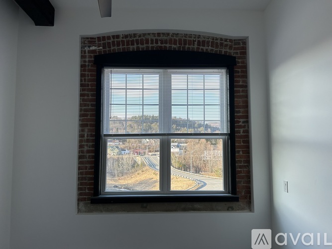 A window with blinds overlooking a road with trees.