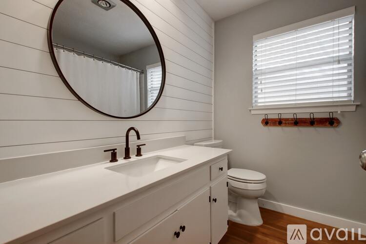 A bathroom with a white sink and toilet, a round mirror, and a window with blinds.