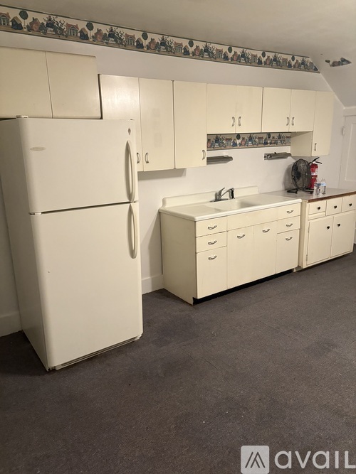 A white refrigerator stands in a kitchen with white cabinets and a white sink.