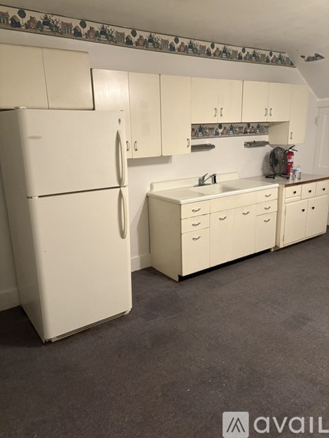 A white refrigerator stands in a kitchen with white cabinets and a white sink.
