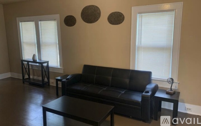 A black leather couch sits in a living room with a coffee table and two windows.