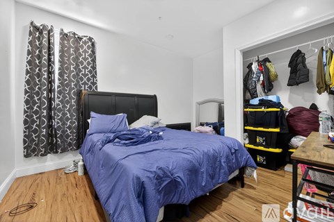 A bedroom with a bed, a wooden table, and a wardrobe with clothes hanging on it.