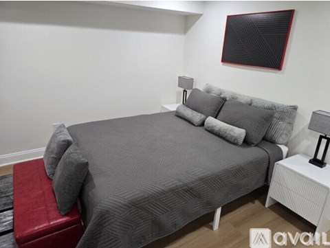 A bedroom with a grey bedspread and pillows, a red bench, and a white nightstand with a lamp.
