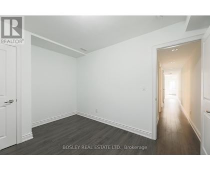 A spacious room with wooden flooring and white walls, with a door on the left and a hallway on the right.