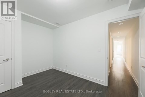 A spacious room with wooden flooring and white walls, with a door on the left and a hallway on the right.