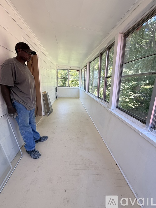 A man in a grey shirt and jeans stands in a room with white walls and windows.