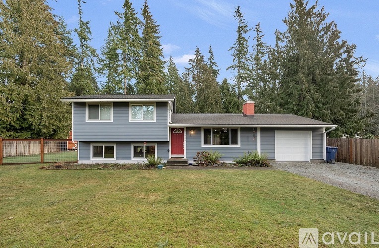 A house with a grey exterior and a red door is surrounded by a grassy area and trees.