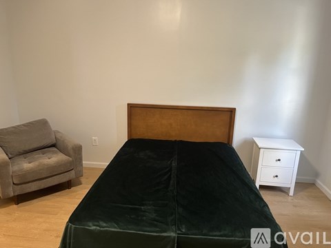A bed with a green cover sits in a room with a grey chair and a small white table.