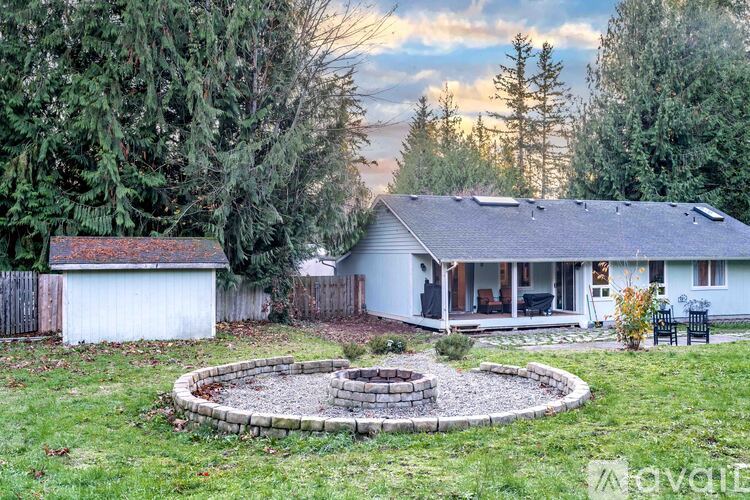A house with a white exterior and a grey roof is surrounded by trees and has a circular fire pit in the front yard.