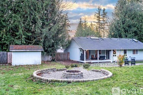 A house with a white exterior and a grey roof is surrounded by trees and has a circular fire pit in the front yard.